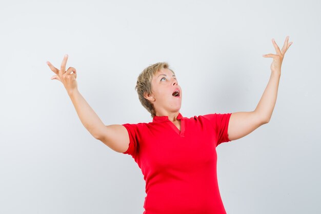 Mature woman looking up, raising hands in red t-shirt and looking astonished