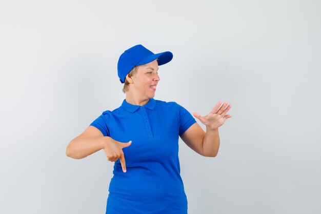 Mature woman pointing down, raising hand in blue t-shirt and looking cheery.