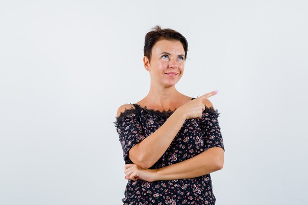 Mature woman pointing right with index finger in floral blouse, black skirt and looking jolly , front view.