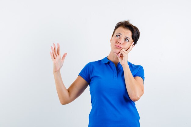 Mature woman raising hand while thinking in blue t-shirt and looking doubtful , front view.