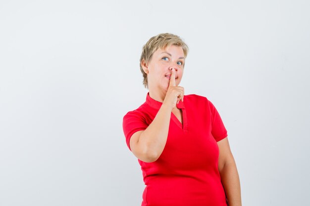 Mature woman showing silence gesture in red t-shirt and looking curious