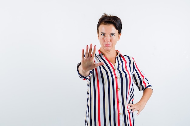 Mature woman showing stop gesture, holding hand on waist in striped blouse and looking serious , front view.