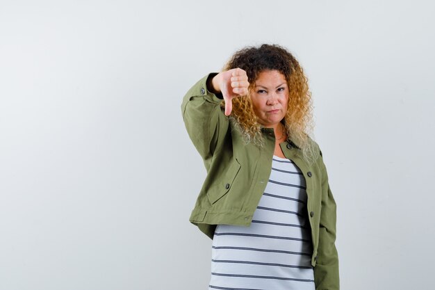 Mature woman showing thumb down in green jacket, t-shirt and looking displeased , front view.