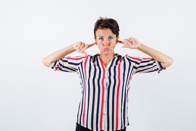 Free photo mature woman in striped shirt plugging ears with index fingers and looking serious , front view.