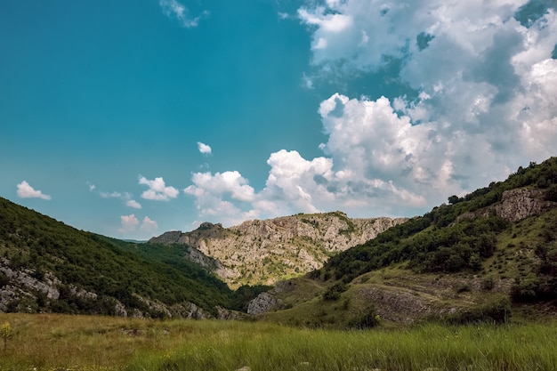 Meadow surrounded by hills covered in bushes and trees under the cloudy sky and sunlight