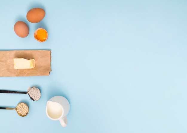 Free photo measuring spoon with oat barn; flour; eggs; butter and milk on blue backdrop