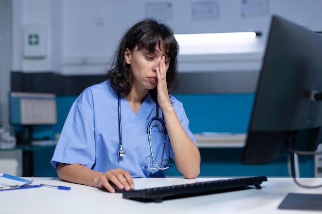 Medical assistant falling asleep while using computer and keyboard for healthcare. Woman nurse using monitor on desk, feeling exhausted and working late at night. Tired practitioner