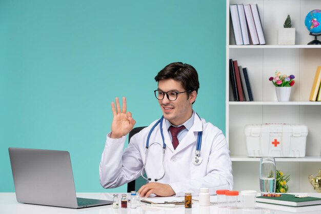 Medical cute handsome doctor in lab coat working remotely on computer showing ok sign