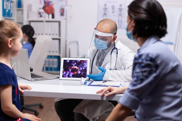 Medical specialist presenting coronavirus using tablet sitting on desk in medical office