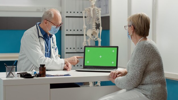 Medical specialist showing greenscreen on laptop to woman at checkup in cabinet. Looking at blank copyspace background with isolated mockup template and chroma key. Tripod shot.