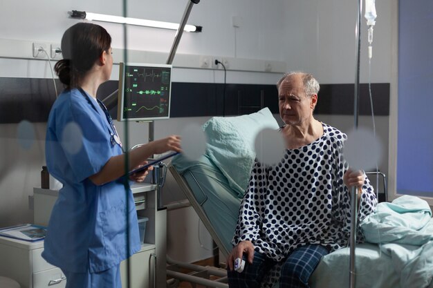 Medical staff with stethoscope questioning sick senior man sitting in bed holding iv drip, with painful expression