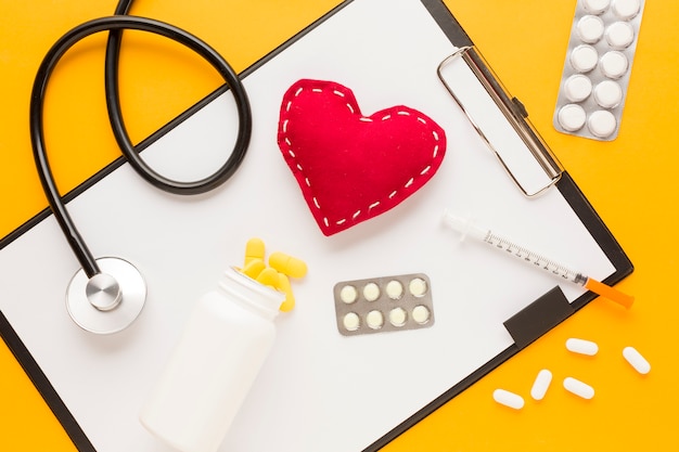 Medicine falling from bottle over clipboard; stethoscope; stitched heart shape; injection; blister packed medicine against yellow desk