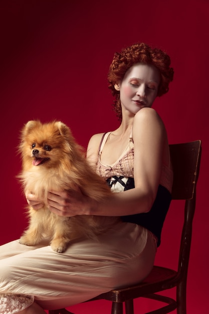 Free Photo medieval redhead young woman as a duchess in black corset and night clothes sitting on a chair on red space with a little puppy or dog