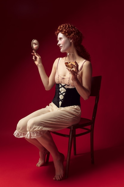 Free photo medieval redhead young woman as a duchess in black corset and night clothes sitting on red space with a mirror and a glass of wine