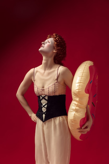 Free photo medieval redhead young woman as a duchess in black corset and night clothes standing on red wall with a swim circle as a donut. concept of comparison of eras, modernity and renaissance.