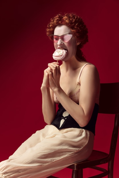 Free photo medieval redhead young woman as a duchess in black corset, sunglasses and night clothes sitting on a chair on red space with a candy