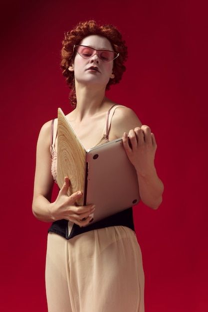 Free photo medieval redhead young woman as a duchess in black corset, sunglasses and night clothes standing on red wall with a laptop as a book. concept of comparison of eras, modernity and renaissance.