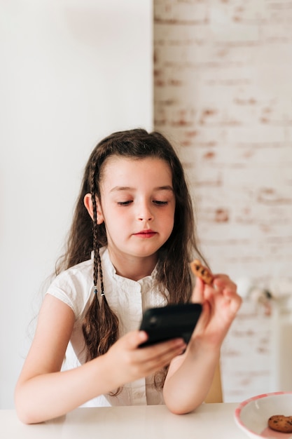 Medium shot girl with phone eating cookies