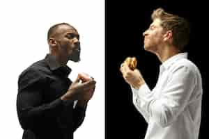 Free photo men eating a hamburger and donut on a black and white background. the happy afro and caucasian men. the burger, fast, unhealthy food concept