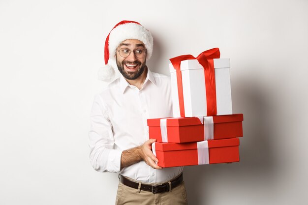 Merry christmas, holidays concept. Excited man celebrating xmas, wearing santa hat and holding gifts, standing  