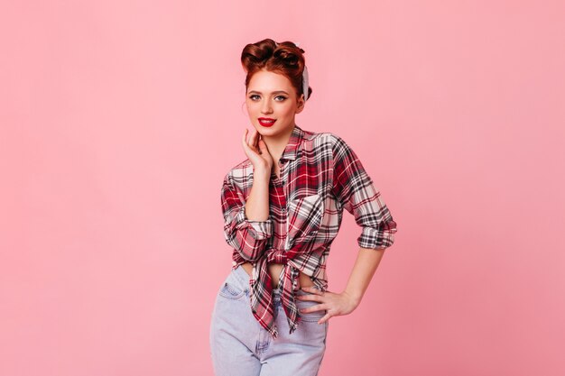 Merry pinup girl smiling on pink space. Studio shot of stylish ginger woman in blue jeans and checkered shirt.