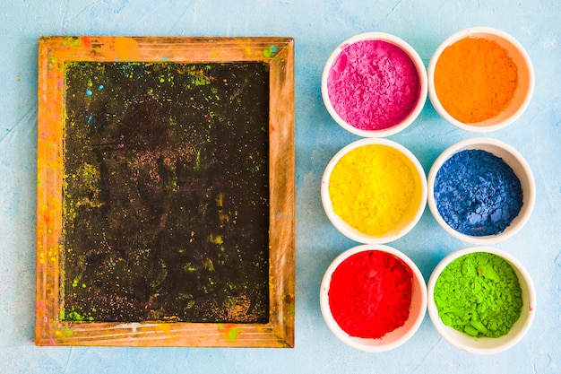 Free photo messy wooden slate with holi color powder in the white bowls on painted backdrop