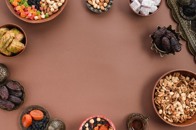 Metallic and earthen bowl of dried fruits; dates; lukum; nuts and baklava arranged on circular shapeover the brown background