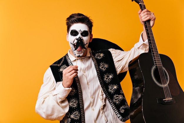 Free photo mexican brown-eyed man with face art in form of skull emotionally shouts, posing with false mustaches and guitar in his hands on orange wall.