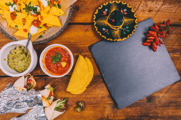 Free Photo mexican hat; wrapped tacos; tasty nachos; salsa sauce; guacamole; black slate and red chilies on table