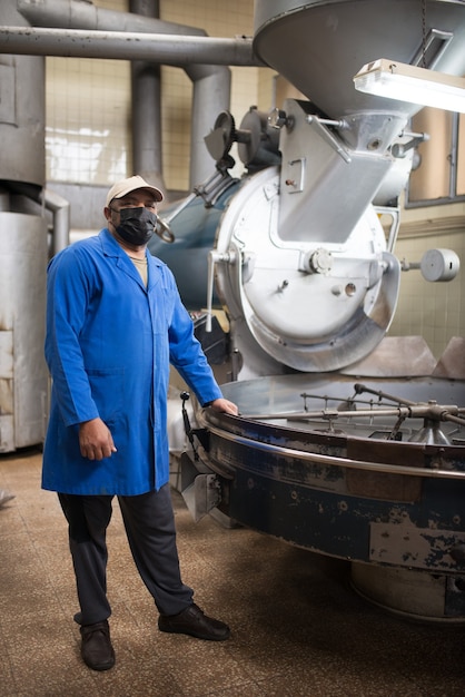 Mid adult coffee roaster standing next to coffee roasting machine. Interior of coffee production workshop with set up roasting equipment