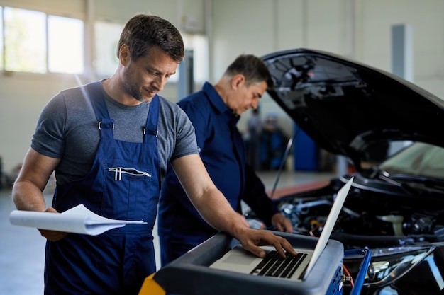 Mid adult mechanic working on laptop while running car diagnostic with his coworker in auto repair shop