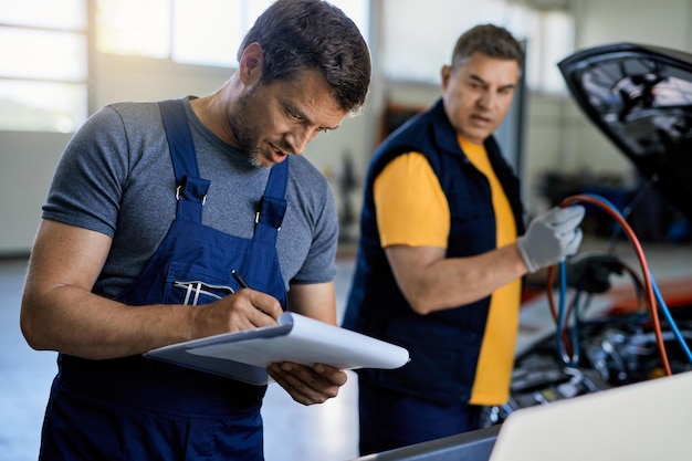 Free Photo mid adult mechanic writing on clipboard while repairing car engine with a coworker in auto repair shop