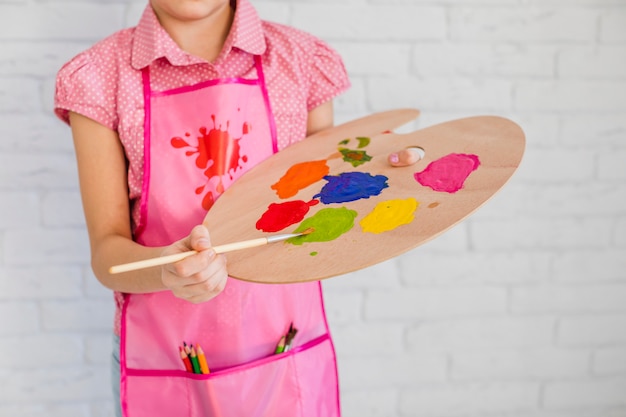 Free photo mid section of a girl wearing pink apron mixing the paint on palette with paintbrush