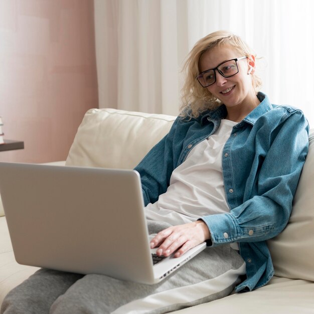 Mid shot blonde woman sitting on couch and working on laptop