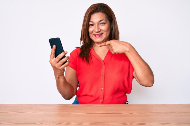 Middle age brunette hispanic woman using smartphone sitting on the table smiling happy pointing with hand and finger