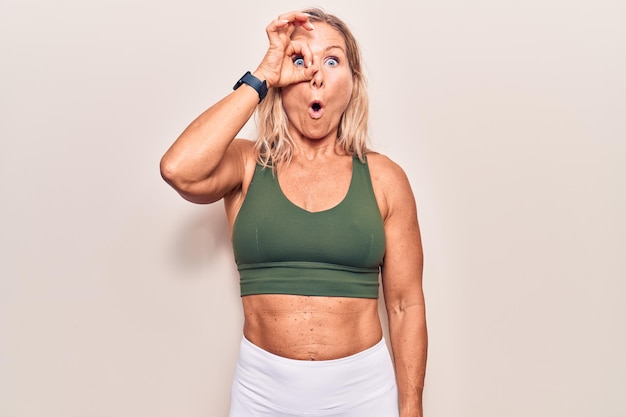 Free photo middle age caucasian blonde woman wearing sportswear doing ok gesture shocked with surprised face, eye looking through fingers. unbelieving expression.