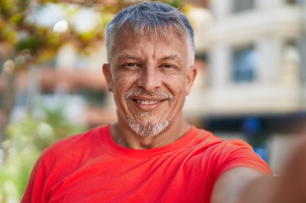 Middle age grey-haired man smiling confident making selfie by camera at park