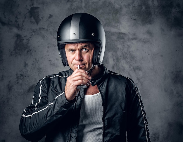 Free Photo middle age male in motorcycle helmet and leather jacket smoking a cigarette on grey background.