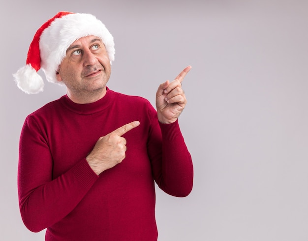 Middle age man wearing christmas santa hat looking up with smile on face pointing with index fingers to the side  standing over white  background