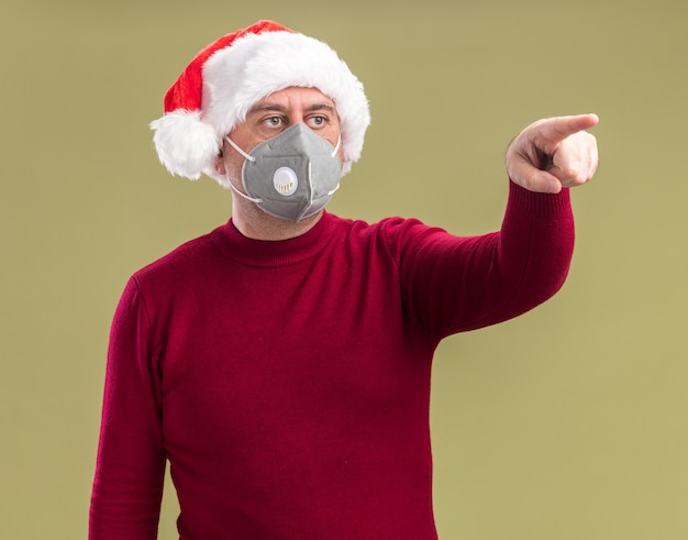 Middle age man wearing christmas santa hat wearing facial protective mask  looking aside with worried expression pointing with index finger to the side standing over green  background