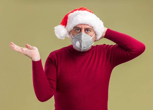 middle age man wearing christmas santa hat wearing facial protective mask looking at camera confused with arm raised standing over green background