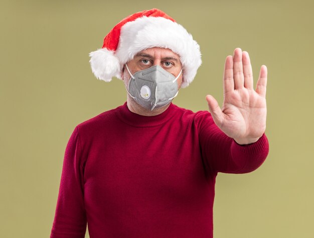 Middle age man wearing christmas santa hat wearing facial protective mask  making stop gesture with hand  with serious face  standing over green  wall