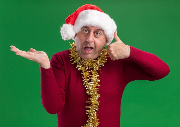 Middle age man wearing christmas santa hat with tinsel around neck   confused showing thumbs up and presenting copy space with arm of his hand standing over green  wall