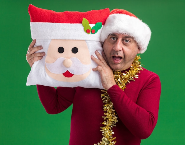 middle age man wearing christmas santa hat with tinsel around neck holding christmas pillow looking at camera amazed and surprised standing over green background