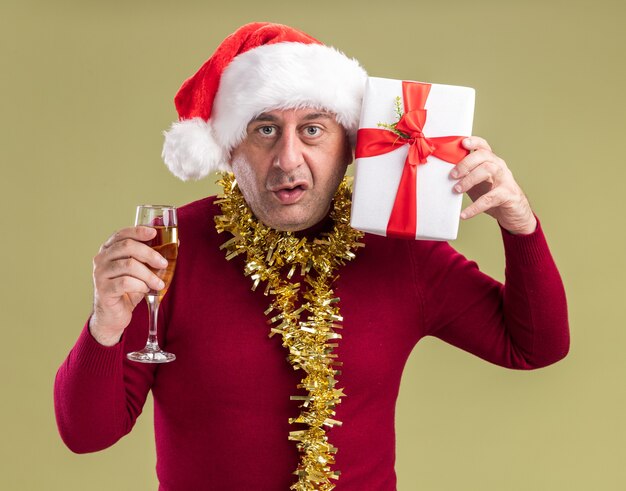 Middle age man wearing christmas santa hat with tinsel around neck holding christmas  present and glass of champagne  looking at camera confused standing over green  background