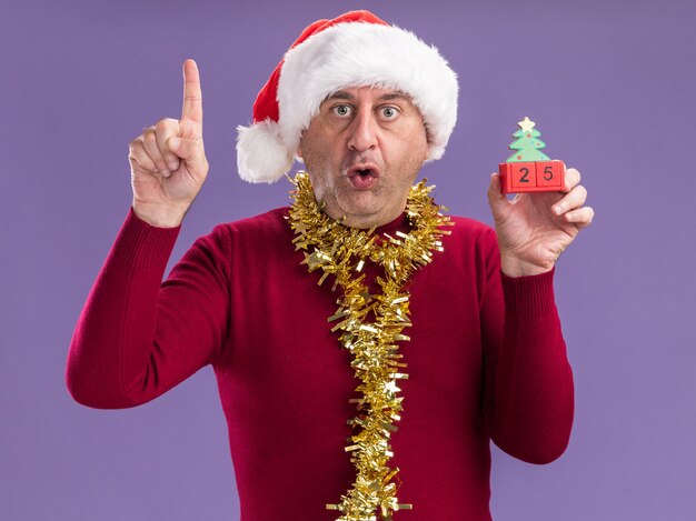 Middle age man wearing christmas santa hat  with tinsel around neck holding toy cubes with date twenty five looking at camera mazed and surprised showing index finger standing over purple background