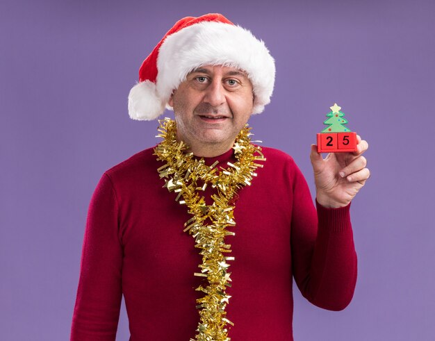 middle age man wearing christmas santa hat with tinsel around neck holding toy cubes with date twenty five looking at camera smiling standing over purple background