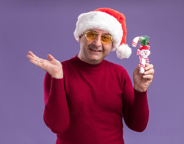 Free photo middle age man wearing christmas santa hat in yellow glasses holding christmas candy cane smiling presenting with arm standing over purple background