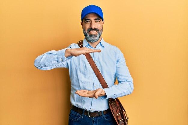 Middle age man with beard and grey hair wearing delivery courier cap gesturing with hands showing big and large size sign, measure symbol. smiling looking at the camera. measuring concept.