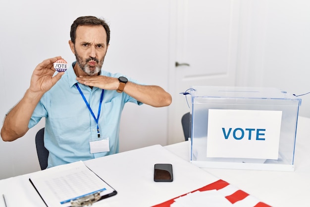 Free photo middle age man with beard sitting by ballot holding i vote badge cutting throat with hand as knife threaten aggression with furious violence